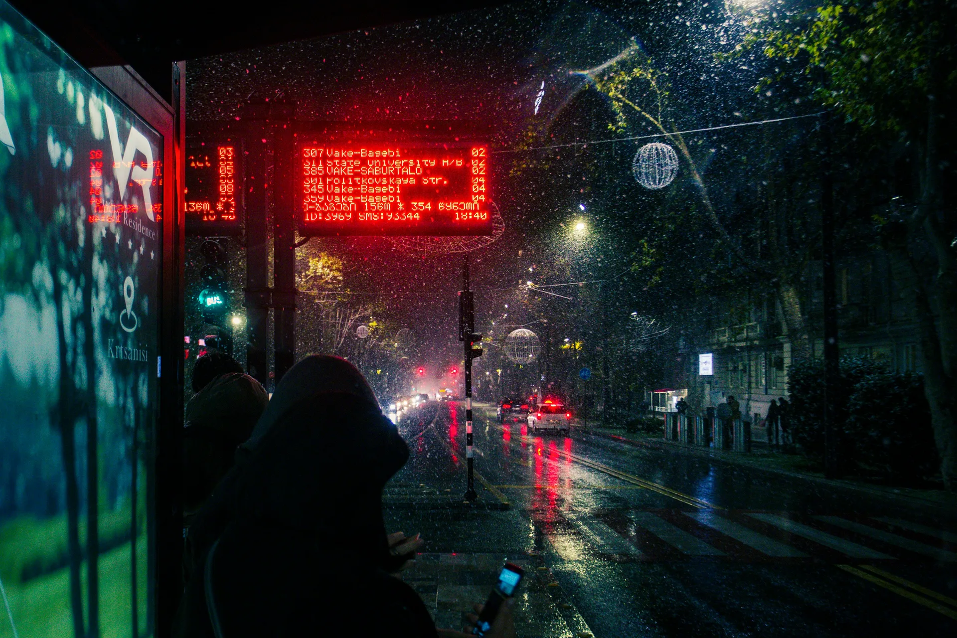 Tbilisi bus stop at night showing LED display with route numbers and rainy street reflections